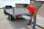 Man in a red shirt lowering a metal ramp from a flatbed trailer at a construction site with a pickup nearby