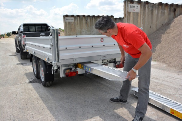 Man in a red shirt lowering a metal ramp from a flatbed trailer at a construction site with a pickup nearby