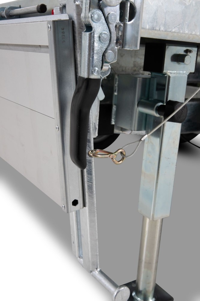 Close-up of a metal gate latch with a black grip, secured by a steel cable and brass ring on an industrial frame.