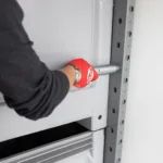 Worker wearing a red glove inserts or removes a metal locking pin into a shelving upright on a gray rack.