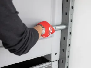 Worker wearing a red glove inserts or removes a metal locking pin into a shelving upright on a gray rack.